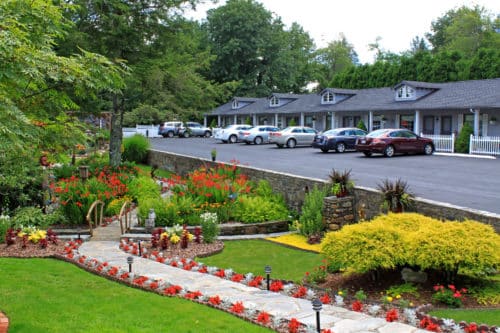 A bright garden stretches in front of a small motor inn with cars parked in front of the room entrances.